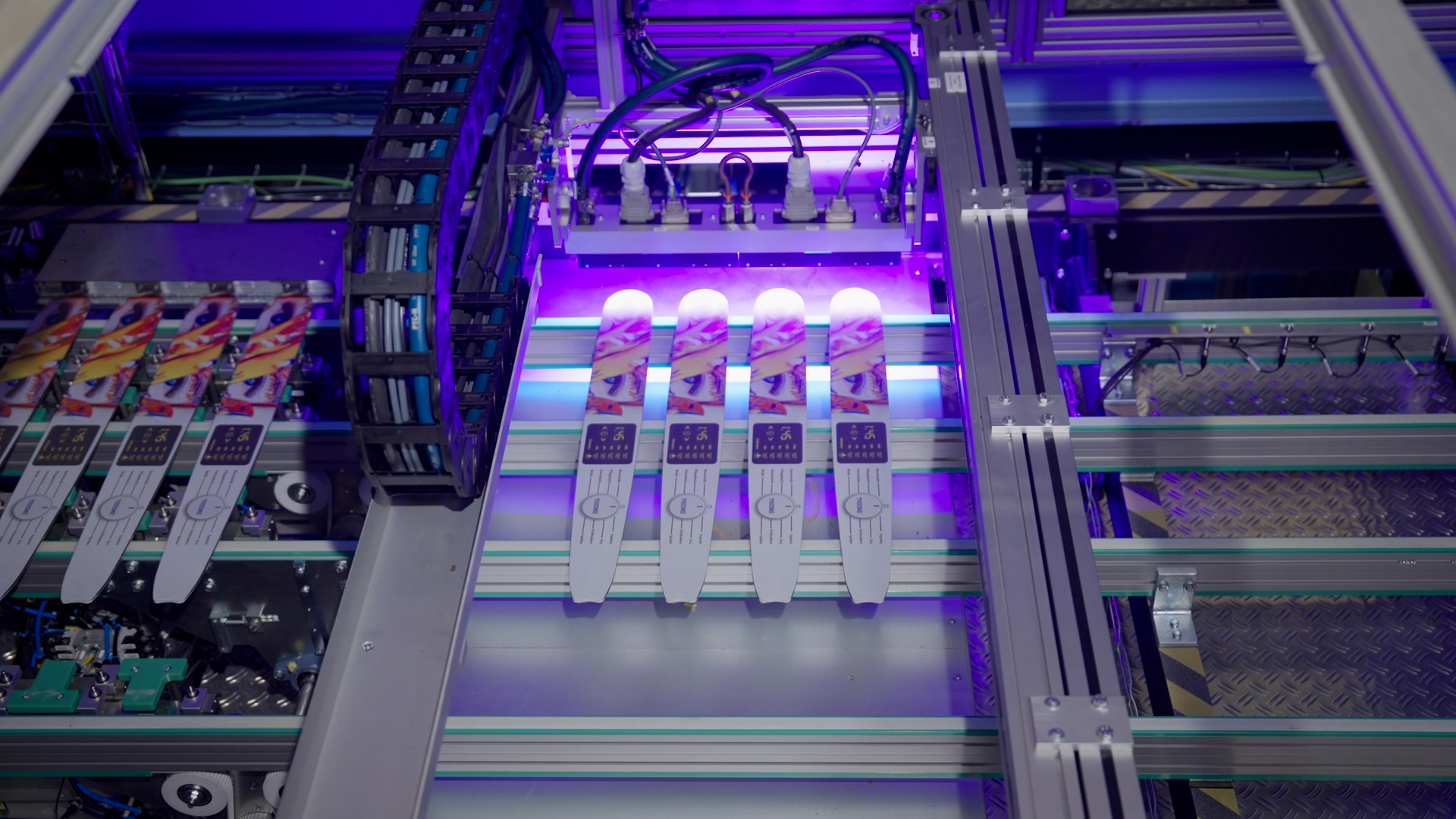 Digitaldruckanlage bedruckt Führungsschienen für Kettensägen; vier Schienen unter UV‑Licht im Druckbereich, Fördertechnik und Kabelkette sichtbar