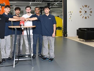 Sechs junge Menschen stehen in Arbeitskleidung um einen Tisch und drücken gemeinsam einen roten Knopf, Symbol für den Start einer Ausbildung.