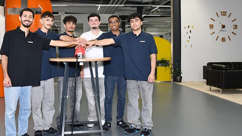 Sechs junge Menschen stehen in Arbeitskleidung um einen Tisch und drücken gemeinsam einen roten Knopf, Symbol für den Start einer Ausbildung.