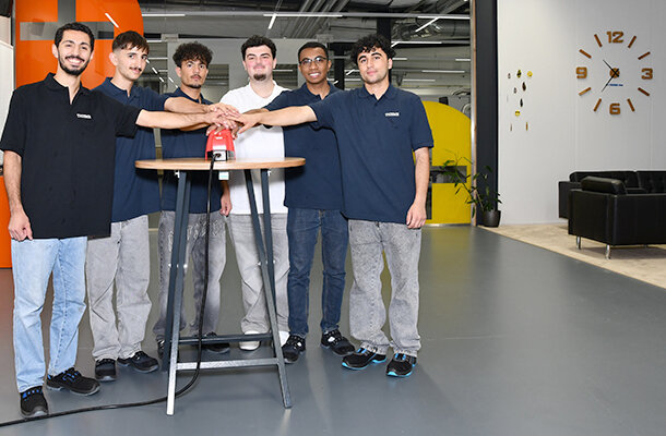 Sechs junge Menschen stehen in Arbeitskleidung um einen Tisch und drücken gemeinsam einen roten Knopf, Symbol für den Start einer Ausbildung.