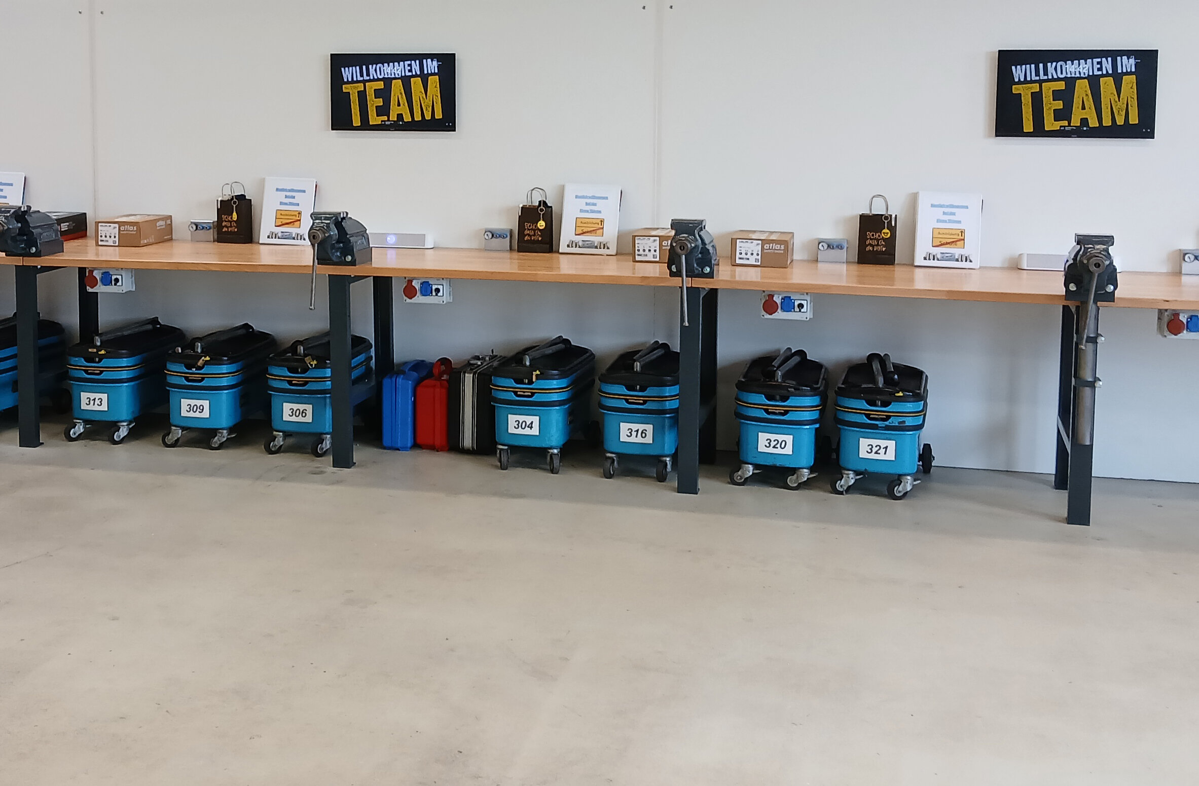 Arbeitsplätze in einem Ausbildungszentrum mit blauen Werkzeugkoffern, Schraubstöcken auf Werkbänken und Schildern mit der Aufschrift „Willkommen im Team“