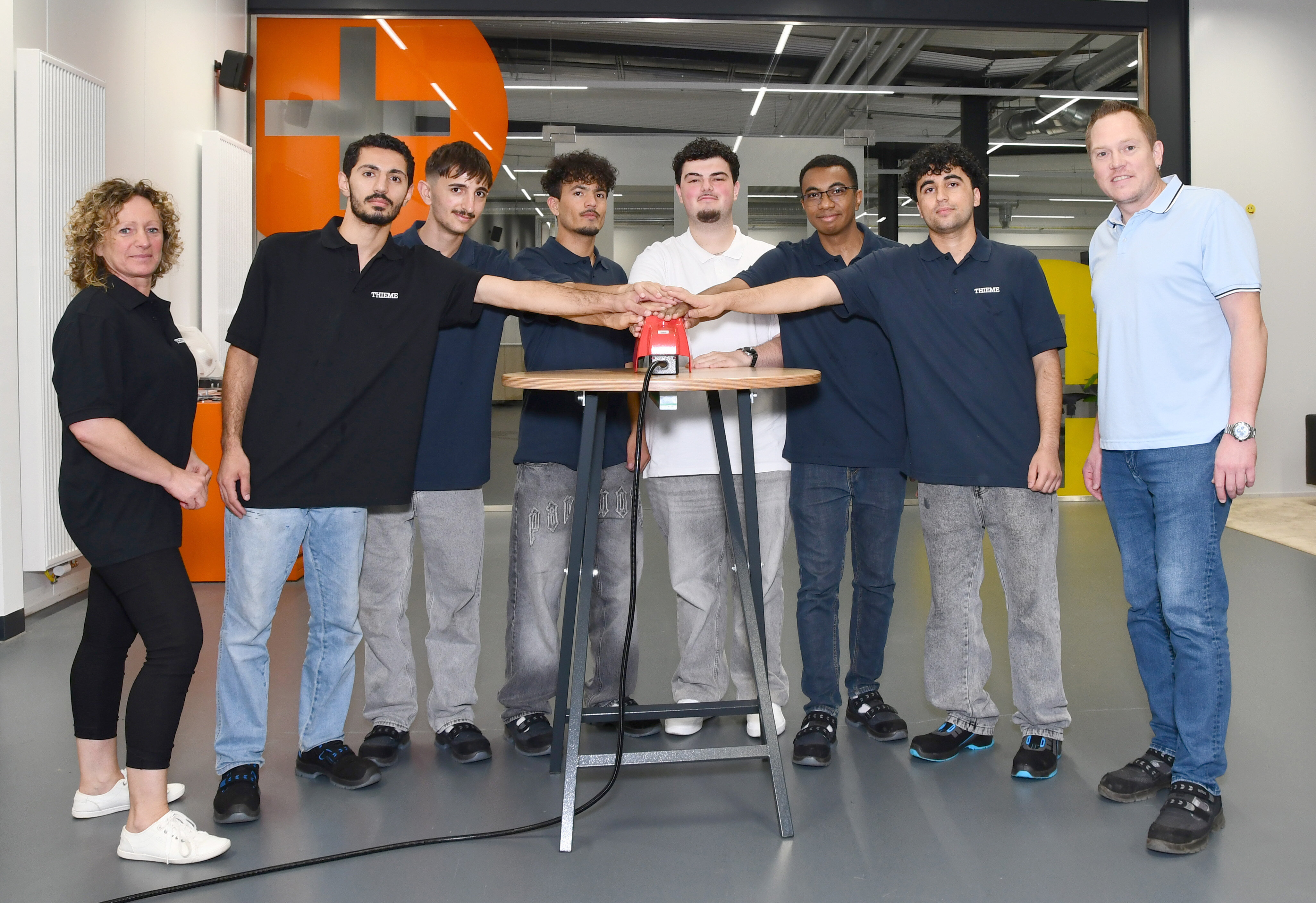 Gruppe von acht Personen steht um einen Tisch und legt gemeinsam die Hände auf einen roten Knopf, Symbol für den Start der Ausbildung.