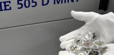 A white-gloved hand holds several colorfully designed collector coins featuring insect motifs in front of a grey machine labeled “505 D MINT.” Visible are coins with bees, beetles, and plant designs.