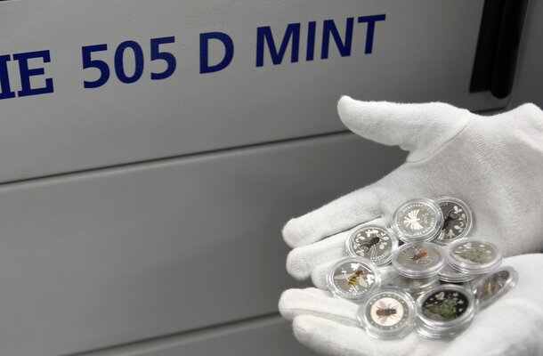 Eine weiße behandschuhte Hand hält mehrere farbig gestaltete Sammlermünzen mit Insektenmotiven vor einer grauen Maschine mit der Aufschrift „505 D MINT“. Zu sehen sind u. a. Münzen mit Bienen, Käfern und Pflanzenmotiven.