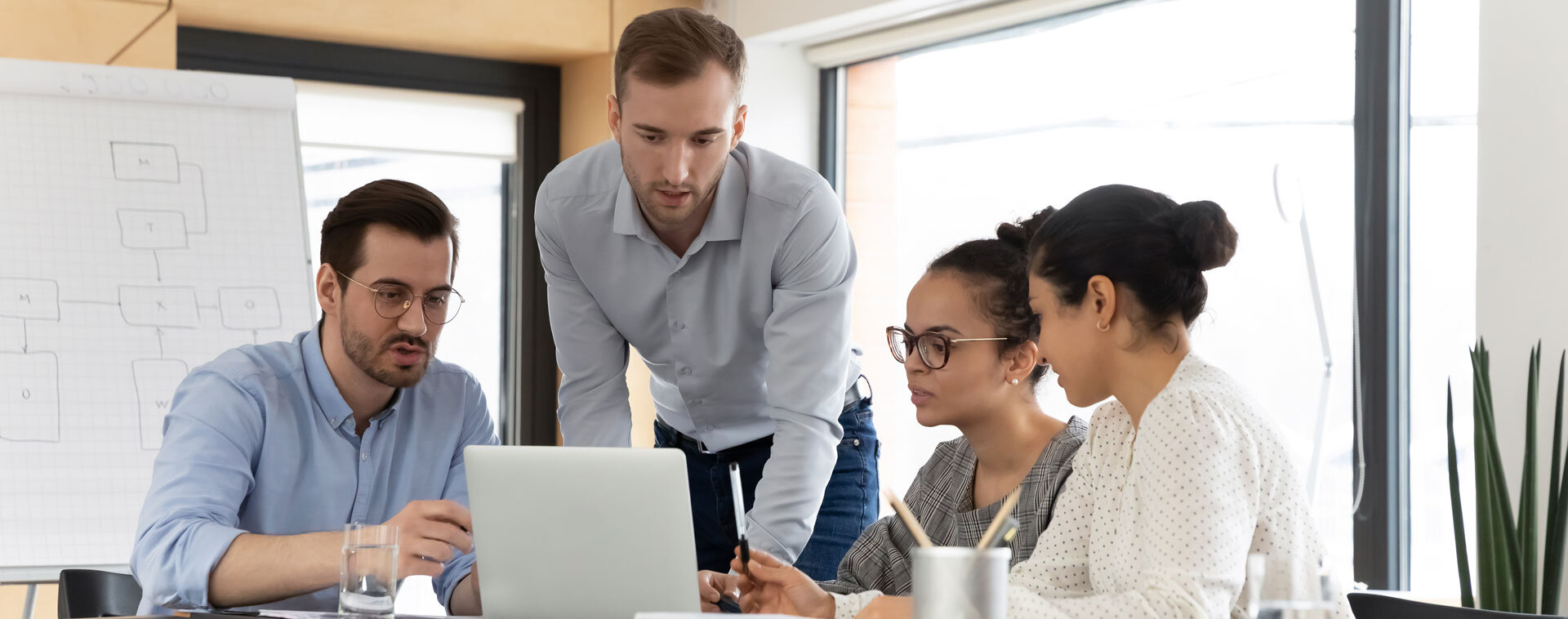 Vier Personen in einem modernen Büro arbeiten gemeinsam am Laptop; eine Person steht über den Tisch gebeugt und erklärt etwas, im Hintergrund ein Flipchart mit Skizzen und große Fenster. | © AdobeStock_436162070