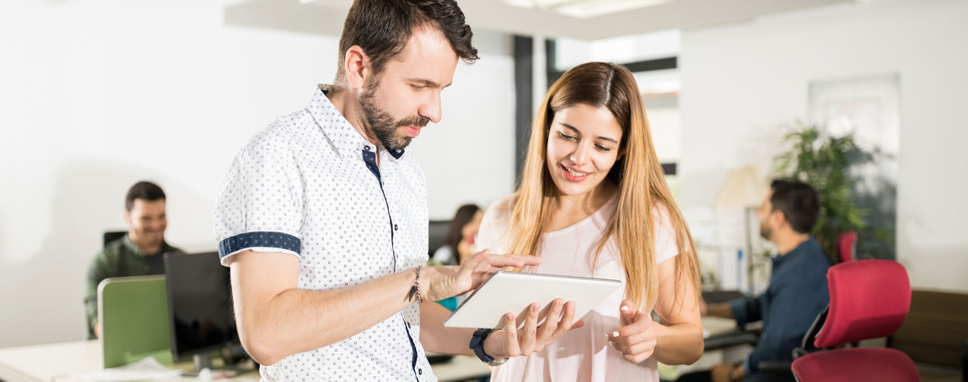 Mann und Frau schauen gemeinsam auf ein Tablet; im Hintergrund sitzen weitere Personen an Schreibtischen | © AdobeStock_211836975